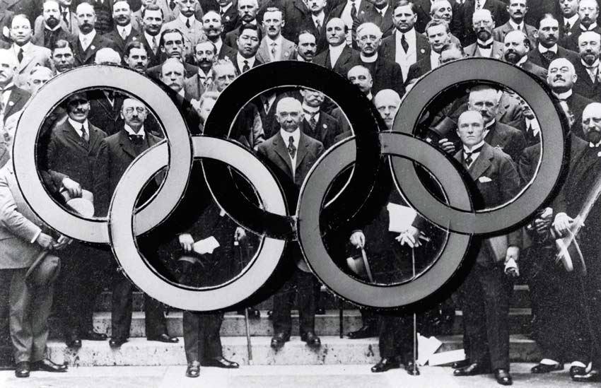 Pierre De Coubertin e il comitato olimpico delle olimpiadi del 1914 dietro i cerchi olimpici. Pierre de Coubertin, Mémoires Olympique, Bureau International de Pédagogie Sportive, Lausanne, 1932. 